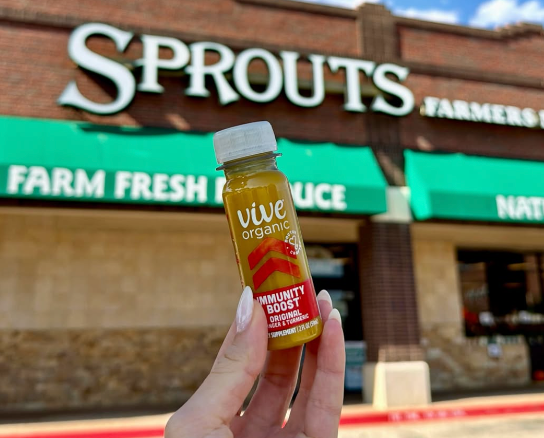 Mão segurando um shot Vive Organic em frente à loja Sprouts Farmers Market.